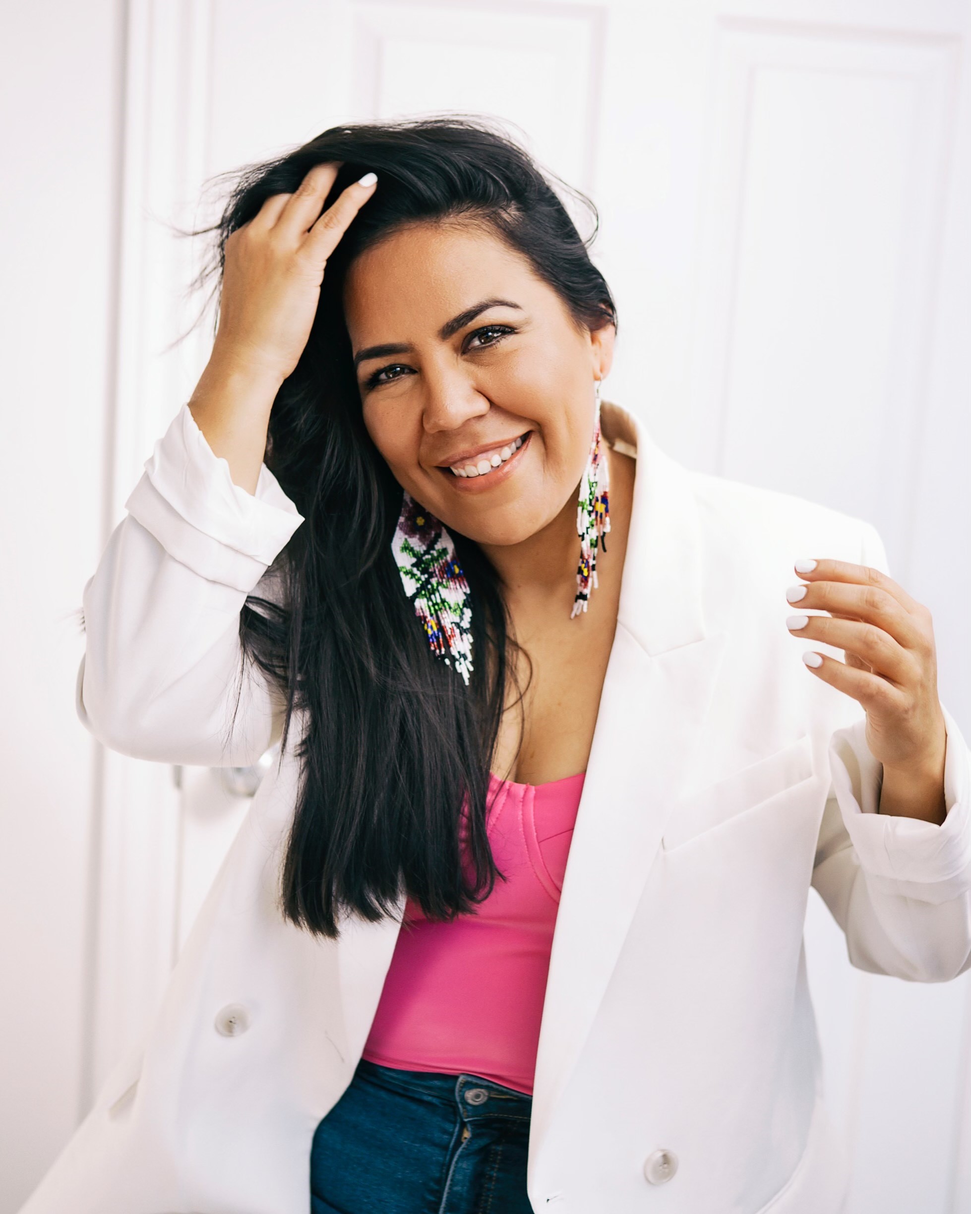 A person with long dark hair smiles, touching their hair. They wear a white blazer, pink top, and colorful beaded earrings, reflecting the vibrant spirit of Indigenous Pathways while standing in front of a light-colored door.